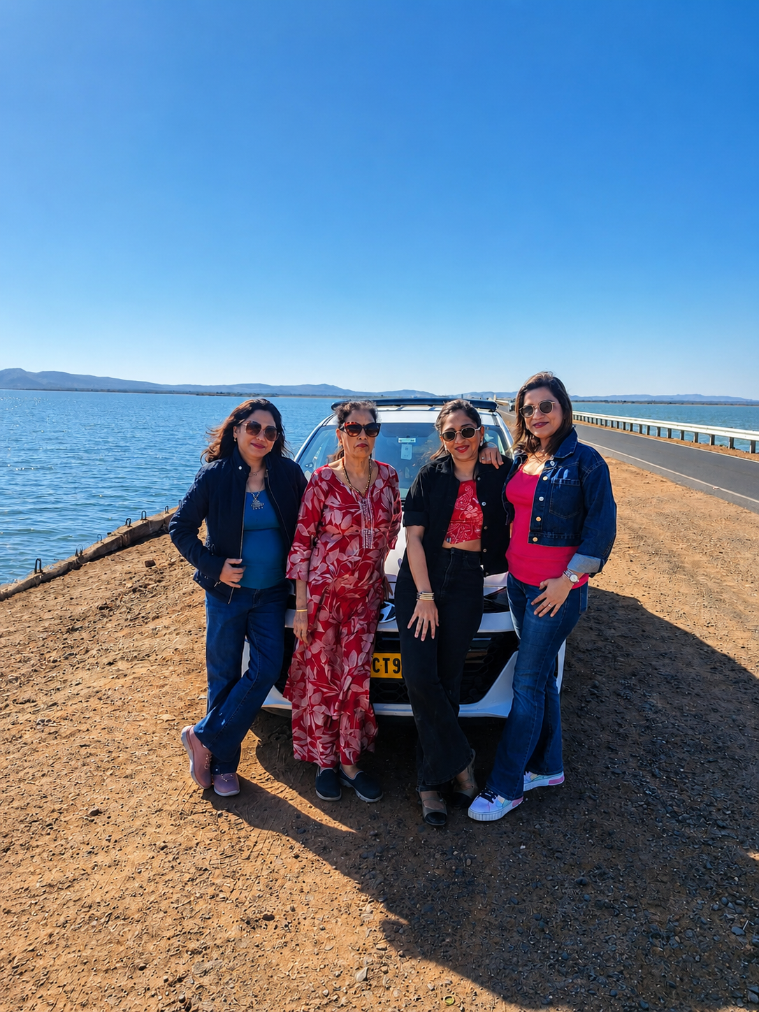 Four women friends standing beside their car on a bridge over water during a Gujarat road trip